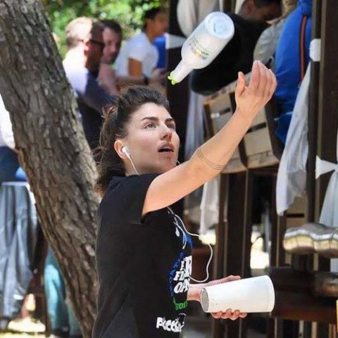 Person juggling two bottles outdoors, wearing earphones, with a crowd in the background.