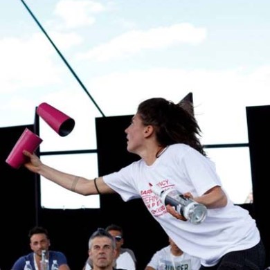 Person juggling shakers, mid-air under a tent roof, with seated spectators watching.