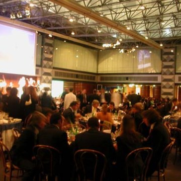 Large banquet hall with people seated at round tables and a stage in the background.