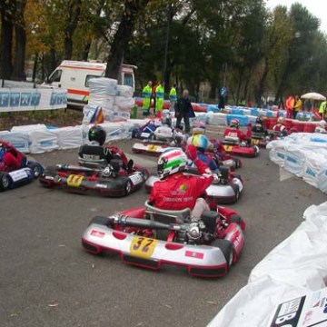 People in helmets racing go-karts on a tree-lined track with barriers.
