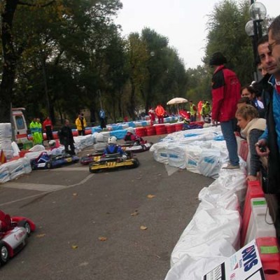 Go-kart race with drivers in helmets on track lined with barriers, spectators watching from the side.