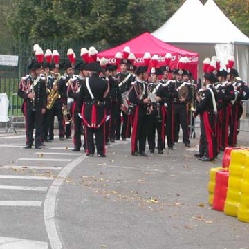 Uniformed marching band with red plumes gathers near tents and barricades outdoors.
