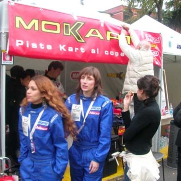 Women in racing suits at a go-kart event with a Mokart sign.