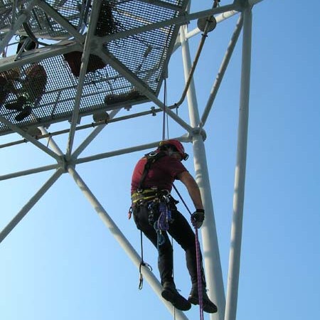 Person in safety gear climbing a metal tower with blue sky background.