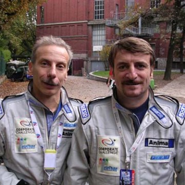 Two men in racing suits with patches pose outdoors on a pathway.