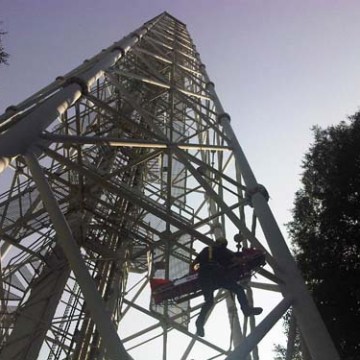 Worker on lift beneath tall metal structure against clear sky.