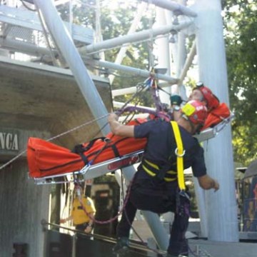 Person in rescue gear operates a stretcher suspended on ropes near a metal structure.