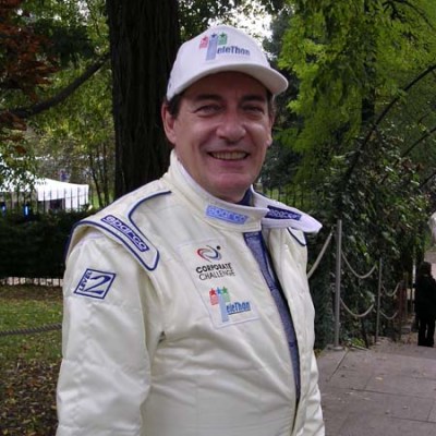 Smiling person in a racing suit and cap, standing outdoors near trees.