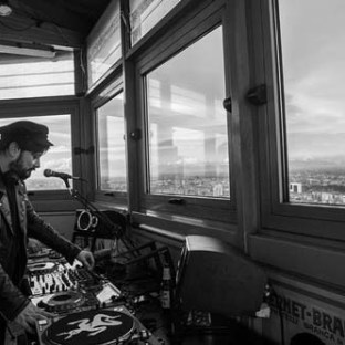 DJ playing music in a booth with panoramic city views through large windows.