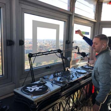 DJ performing in a room with large windows, overlooking a cityscape.
