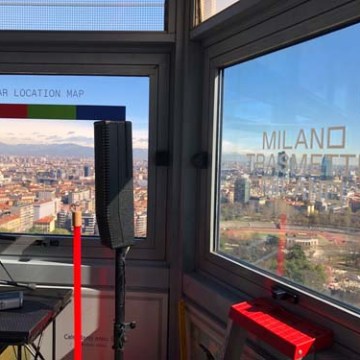 City view through windows with 'Milano Trasmette' text and a solar location map inside room.