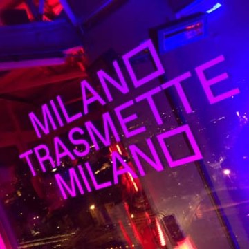 Neon sign with text 'MILANO TRASMETTE MILANO' in pink and purple lights.