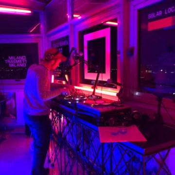 A DJ in a studio with neon pink and blue lights, mixing music on turntables.