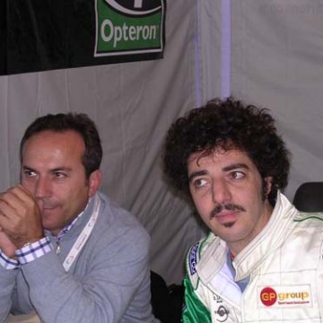 Two men seated indoors, one in a racing suit and the other in a sweater.