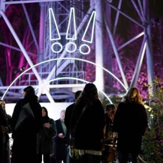 People gather under glowing exclamation marks and arches at night, with trees lit in purple behind.