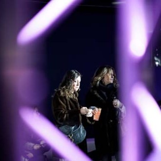 Two women in coats viewed through blurred purple lights indoors.