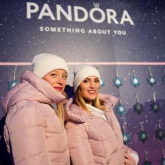 Two women in pink coats and white hats in front of Pandora display with blue stars.