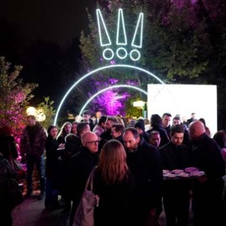 Nighttime outdoor gathering with neon exclamation marks; people mingling under colored lights.
