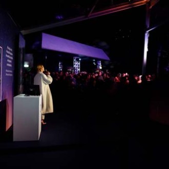Person in white standing on stage at an event with audience in dim lighting.