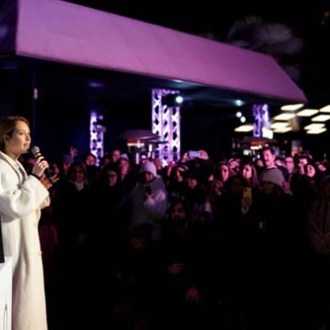 Person in a white coat speaking to a crowd at a nighttime outdoor event.