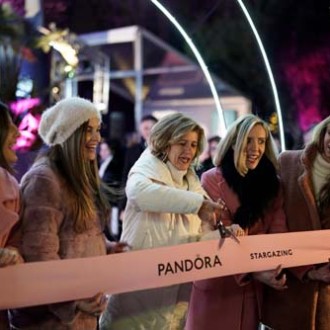 Six women in winter outfits cut a pink ribbon labeled 'PANDORA STARGAZING' at an event.