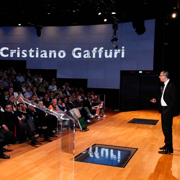 Person speaking on stage to audience, large screen displays 'Cristiano Gaffuri'.
