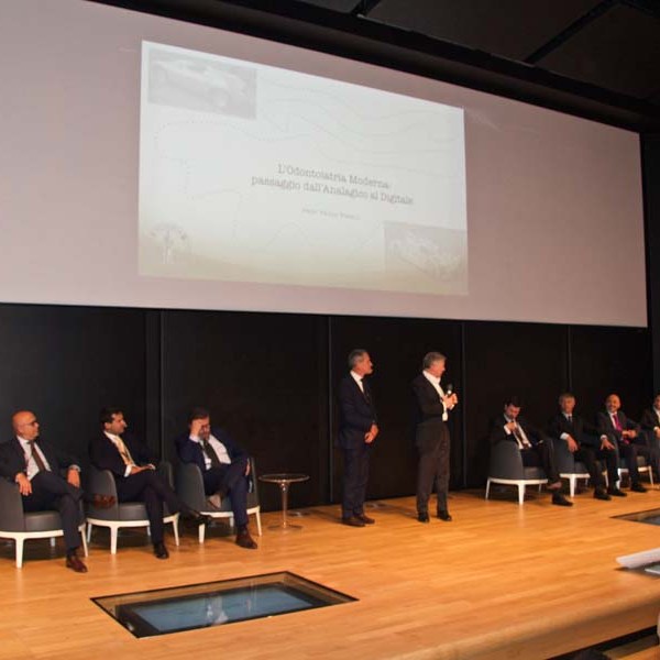 A panel of men in suits on stage with a presentation screen behind them.