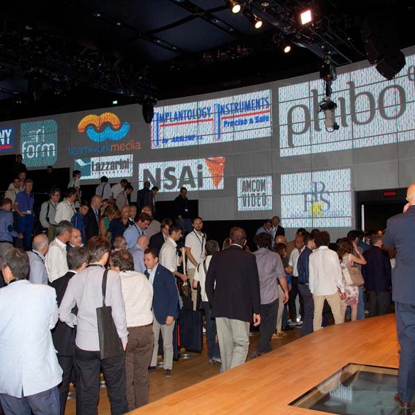 People gathered in a conference hall with logos projected on a large screen.