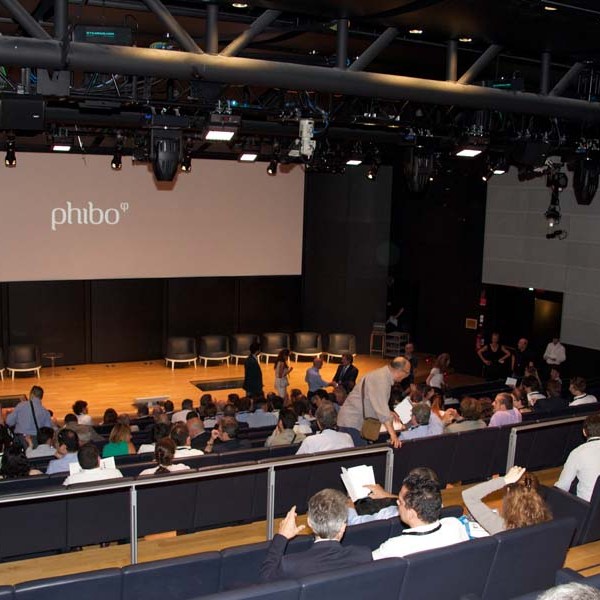 Large auditorium with people seated, facing stage and 'phibo' logo on screen.