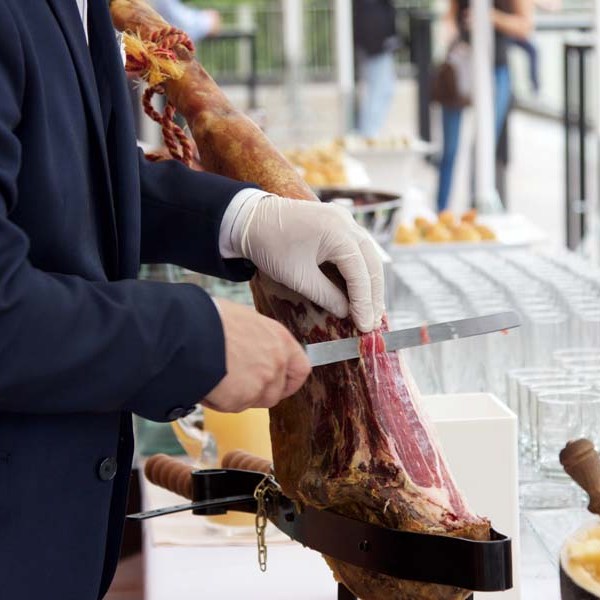 Person slicing cured ham at an event, with glassware in the background.