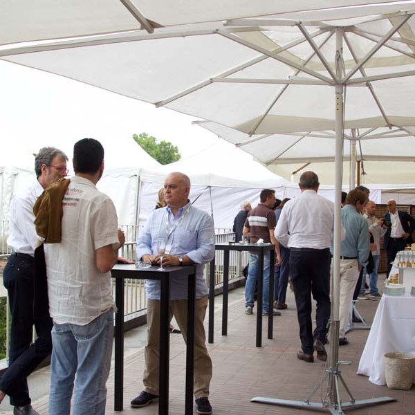 People mingling under umbrellas at an outdoor event with tables and buffet setup.