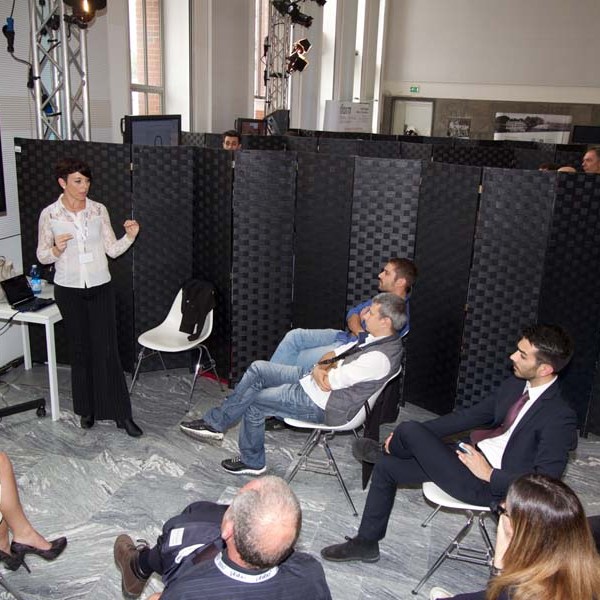 A woman presents data on a screen to a seated audience in a room with black panel dividers.