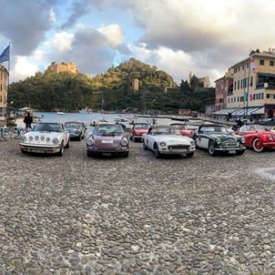Vintage cars lined up on a cobblestone street with colorful buildings and a few people around.