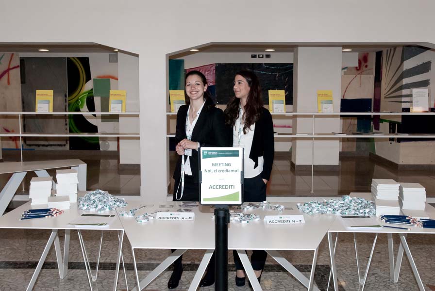 Two women standing behind a conference registration table with sign 'Meeting Noi, ci crediamo.'