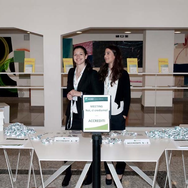 Two women standing behind a conference registration table with sign 'Meeting Noi, ci crediamo.'