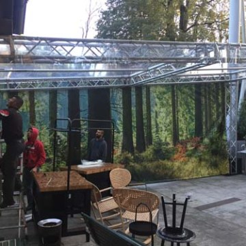 People assembling a clear-roofed structure with a forest backdrop and chairs on the patio.