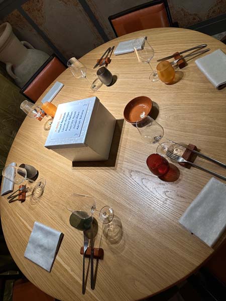 Round wooden table set for six with glasses, napkins, and a centerpiece box.