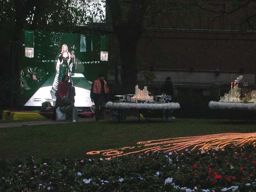 Outdoor scene with a large screen showing a woman on a runway, surrounded by flowers and decorative lights.